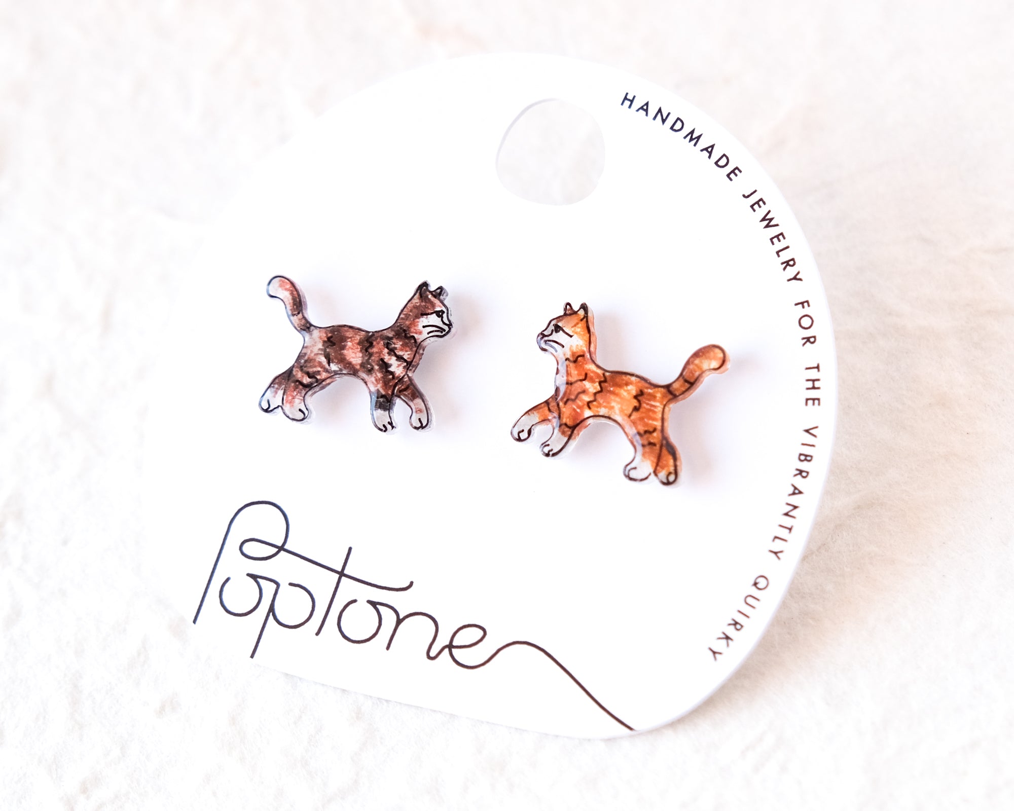 a pair of earrings in the shape of cats, with the packaging for the earrings featuring the brand name 'Poptone' and the description of the earrings as "handmade jewelry for the vibrantly quirky".