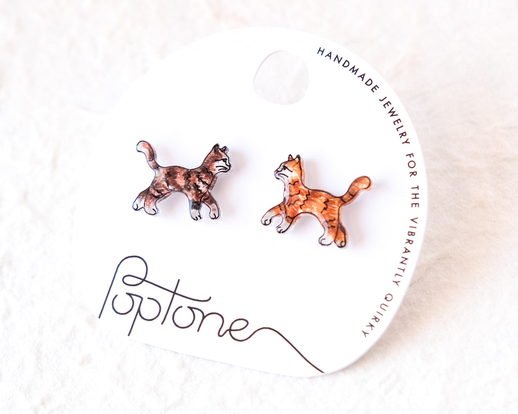 a pair of earrings in the shape of cats, with the packaging for the earrings featuring the brand name 'Poptone' and the description of the earrings as "handmade jewelry for the vibrantly quirky".