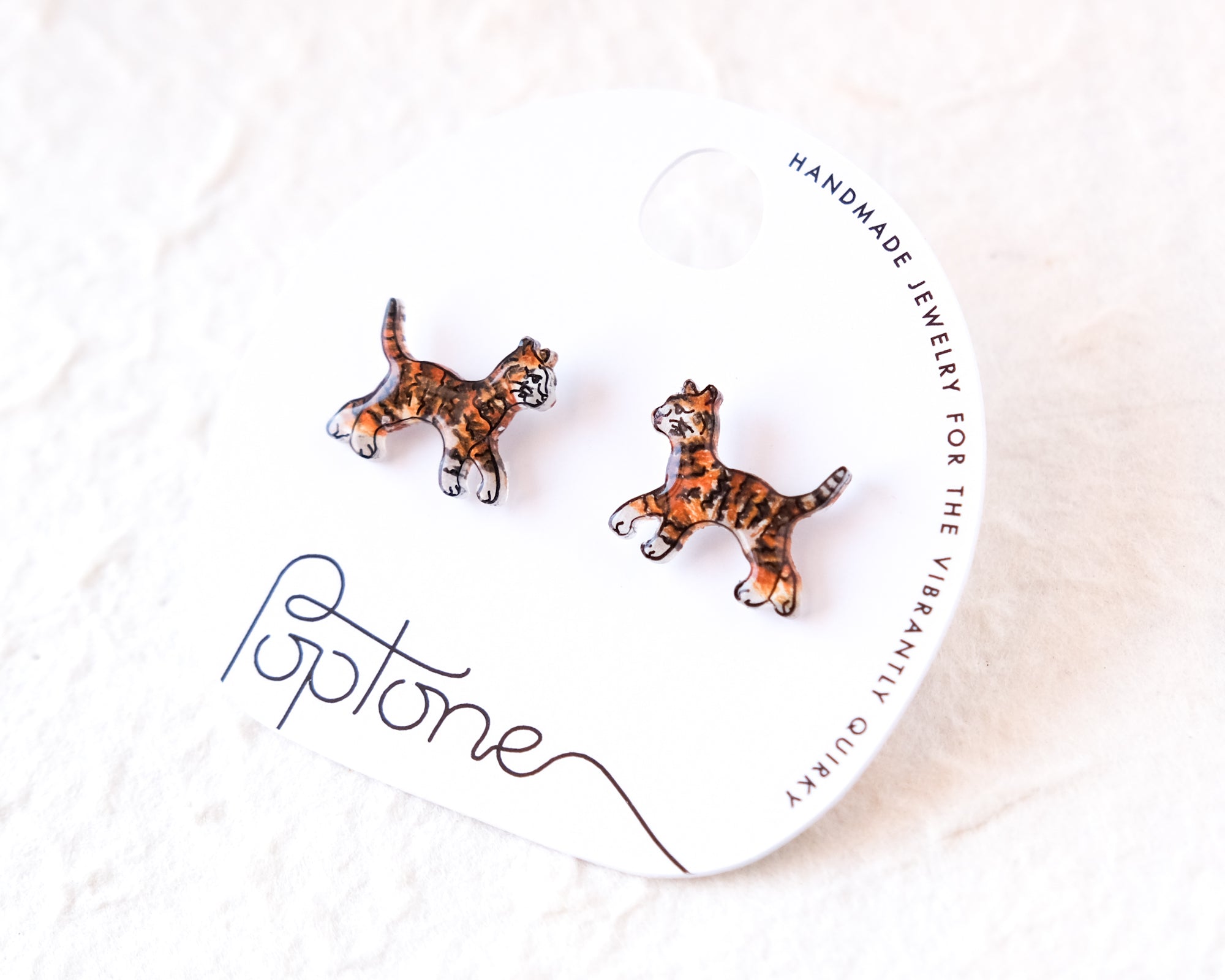 A pair of earrings in the shape of tiger cats, with the packaging for the earrings labeled as "Handmade Jewelry for the Vibrantly Uniquely Beautiful".