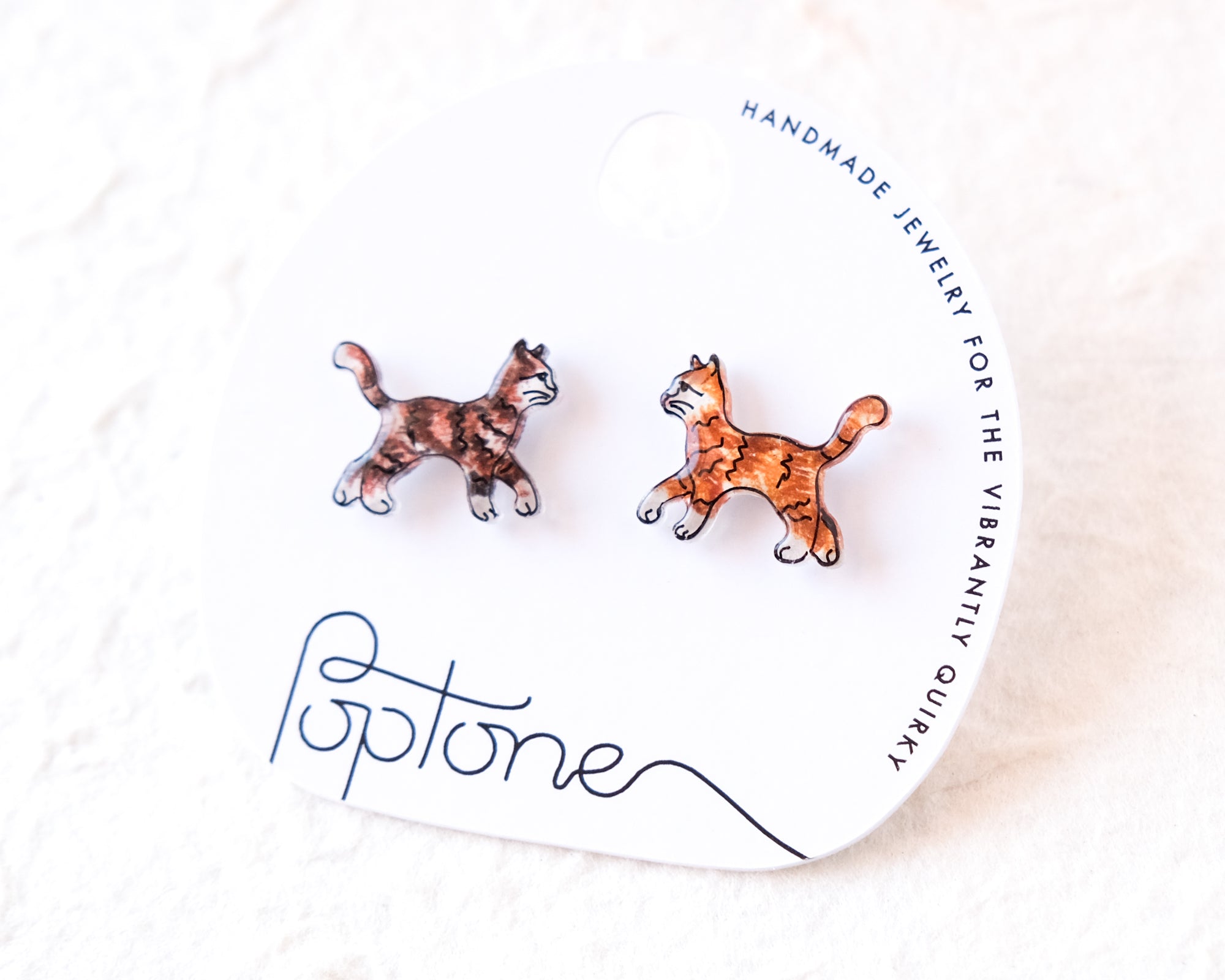 A pair of earrings in the shape of cats, with one cat being brown and the other orange, are displayed on a white background.