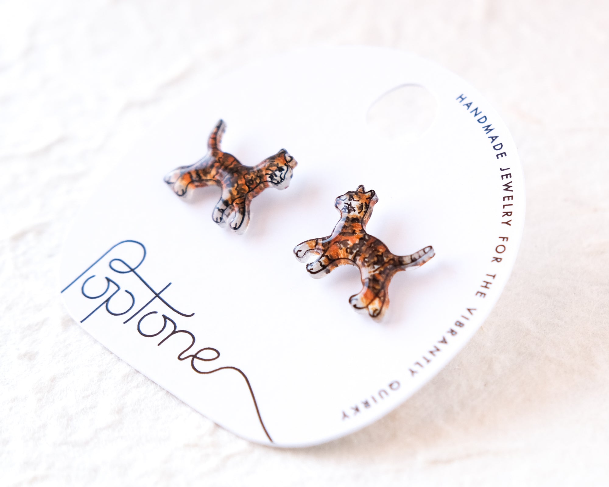 A pair of earrings in the shape of small cats, with the brand name "Poptone" written on the packaging.