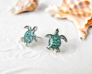 a pair of sea turtle earrings sitting on top of a sandy beach