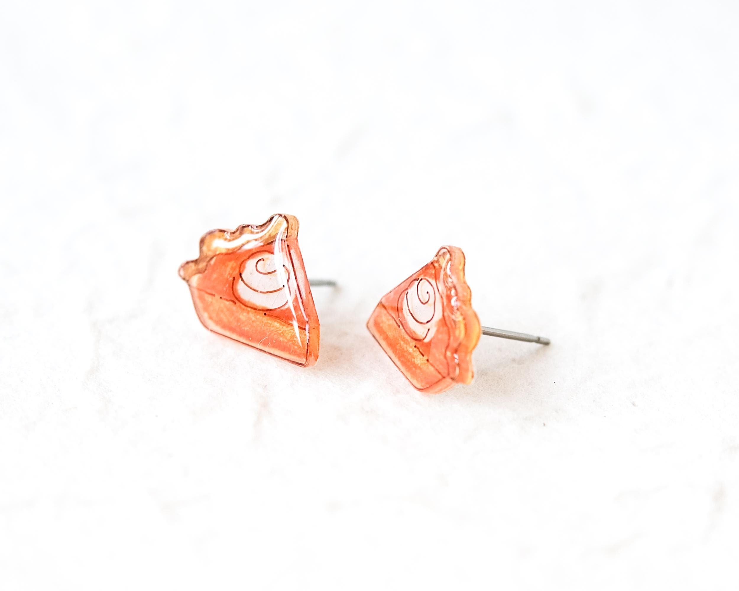 A pair of small, orange-colored earrings shaped like slices of pie or cake, with a white swirl design on the surface.