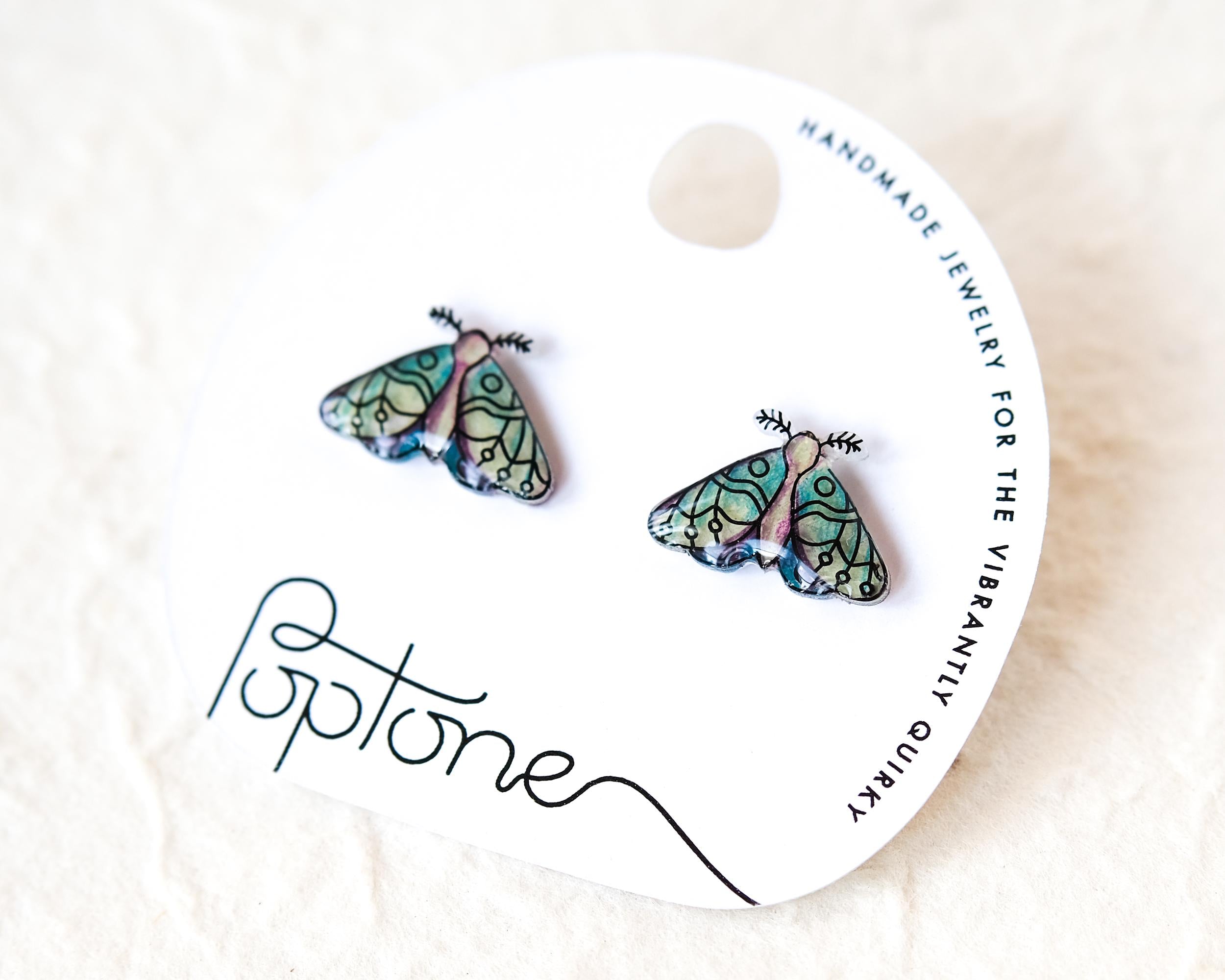 a pair of earrings with butterfly designs, placed on a white circular card with text and logos.