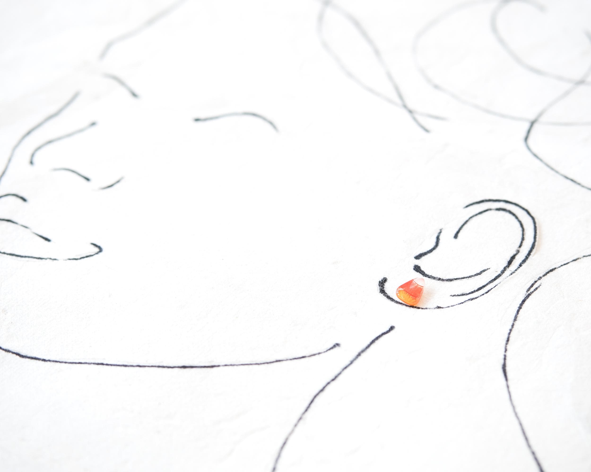 The image is a simple line drawing of a face, with a small orange gemstone placed on the ear.