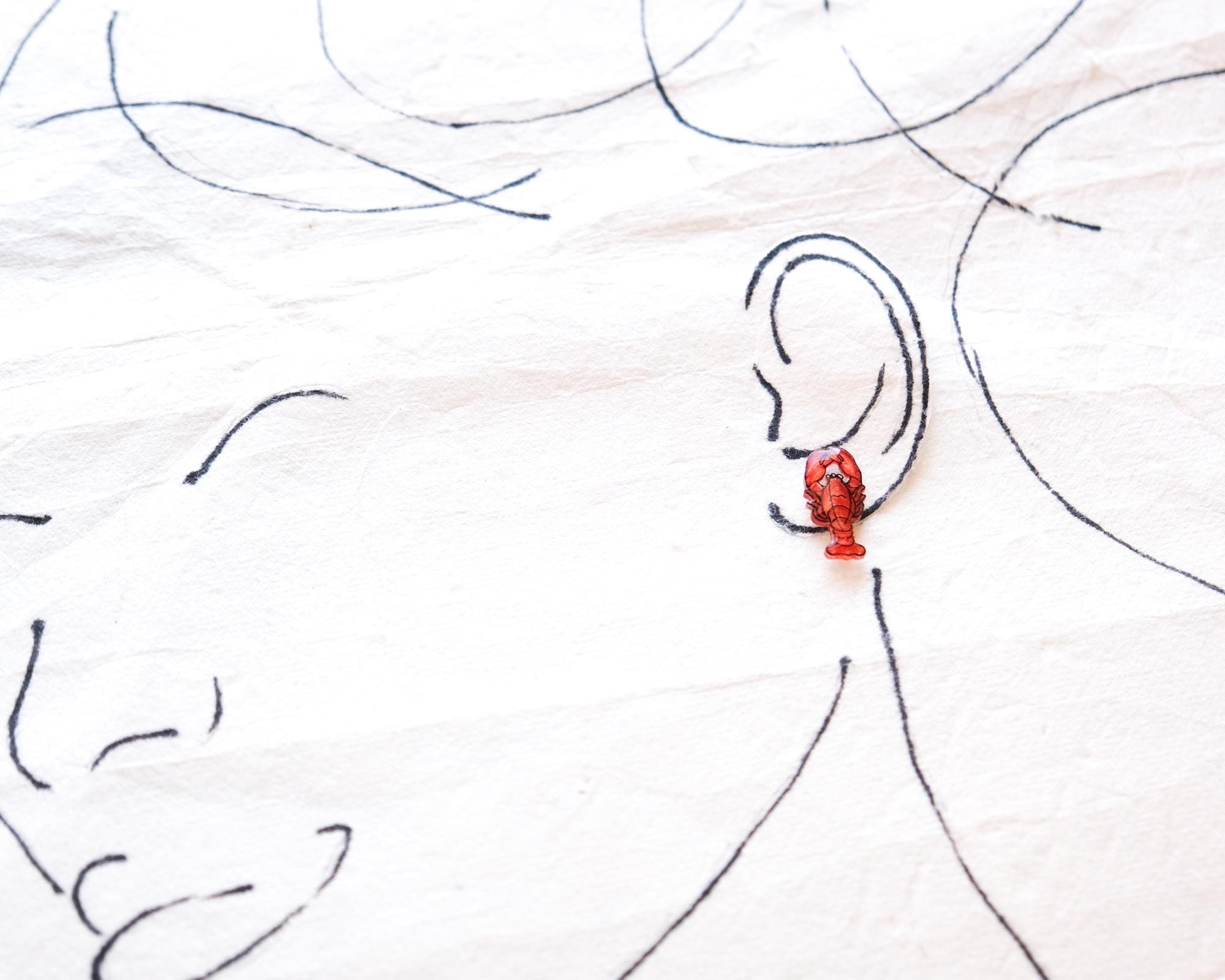 a red lobster earring on a person's ear, with a drawing of a face in the background.
